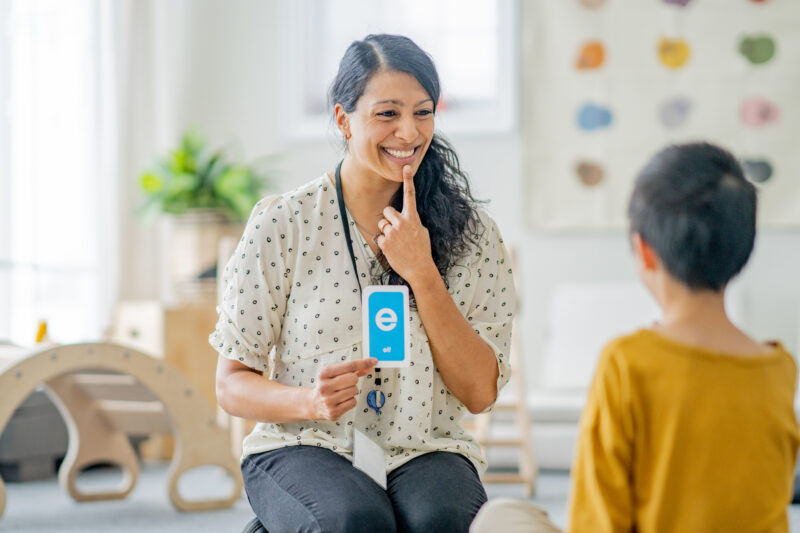 A female speech therapist works with a young boy on his speech as they sit together on the floor.  She is holding up alphabetical cue cards to help as they work through the sounds together.