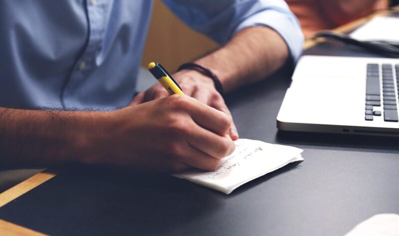 man writing on notebook with laptop in background 