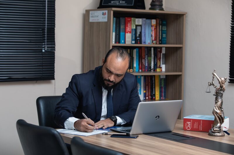 man writing on pad of paper with computer on desk 