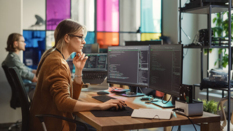 Woman Writes Lines of Code on Desktop PC with Multiple Displays and a Laptop in Stylish Office. Professional Female Data Scientist Using Software To Analyze Information from Internet.