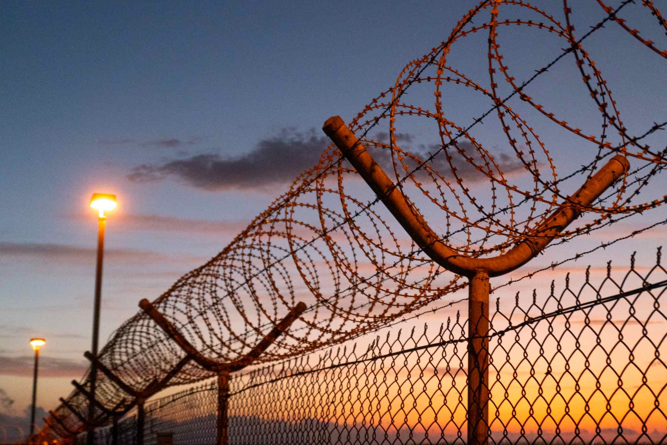 Prompts: Razor Wire Razor wire, barbed wire, mesh wire fencing and floodlights forming part of the security perimeter of Sydney Kingsford Smith Airport. This image was taken from near Shep's Mound, Ross Smith Avenue, Mascot on a hot and sunny evening after sunset