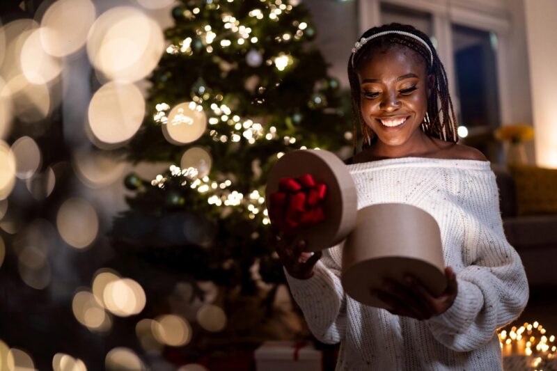Prompt: The Present A young black woman opens a present with a lighted tree behind her