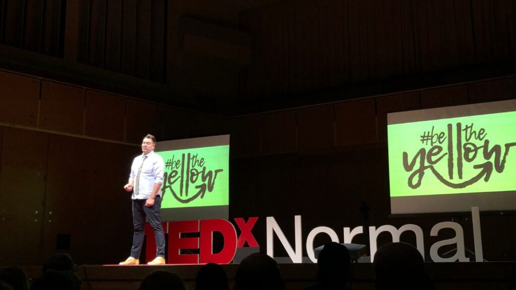 Adam Peterson on TEDx Stage with the words Be the Yellow on screens.